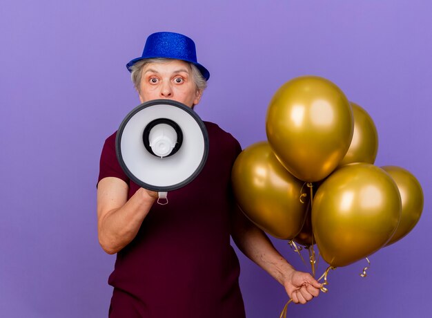 Anciana sorprendida con sombrero de fiesta sostiene globos de helio y altavoz aislado en la pared púrpura con espacio de copia