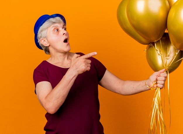 Anciana sorprendida con sombrero de fiesta sostiene globos de helio mirando y apuntando al lado aislado en la pared naranja con espacio de copia