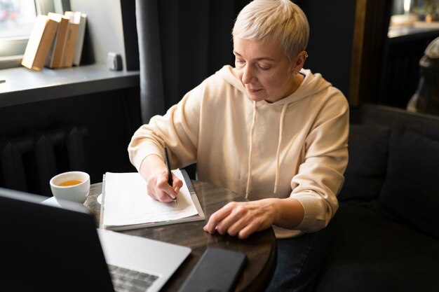 Anciana tomando café en un café mientras trabaja en su computadora portátil y escribe en su cuaderno