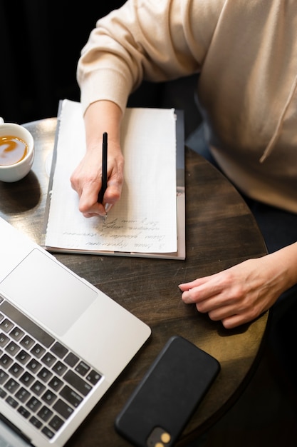 Anciana tomando café en un café mientras trabaja en su computadora portátil y escribe en su cuaderno