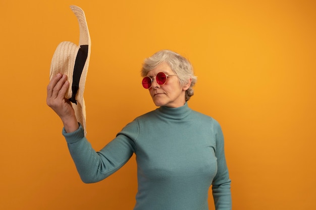 Anciana vistiendo un suéter de cuello alto azul y gafas de sol sosteniendo y mirando el sombrero