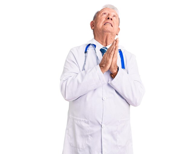 Un anciano apuesto de pelo gris con abrigo médico y estetoscopio rogando y rezando con las manos juntas con expresión de esperanza en la cara muy emocional y preocupada. mendicidad.