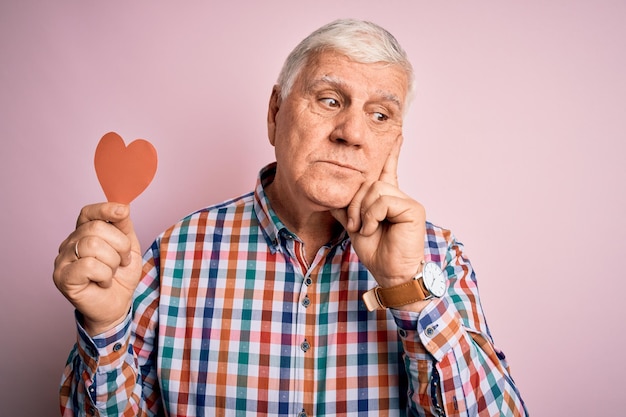 Foto gratuita un anciano apuesto y romántico hombre sosteniendo papel rojo en forma de corazón sobre fondo rosa cara seria pensando en la pregunta idea muy confusa