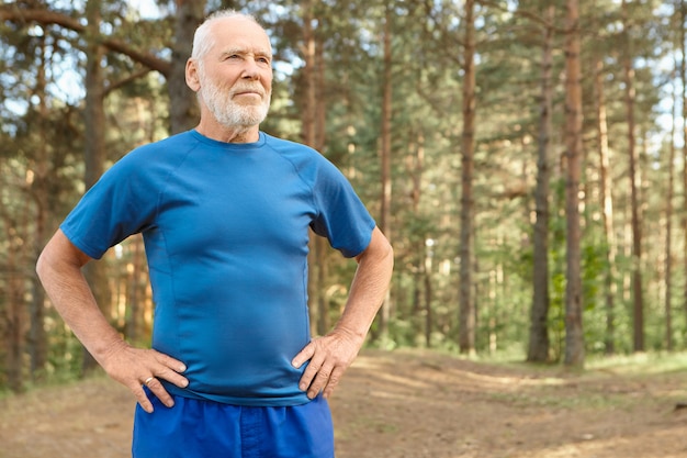 Anciano autodeterminado en entrenamiento de jubilación al aire libre en madera de pino, cogidos de la mano en la cintura, haciendo ejercicios para calentar el cuerpo antes de correr. Hombre jubilado barbudo recuperando el aliento después del entrenamiento