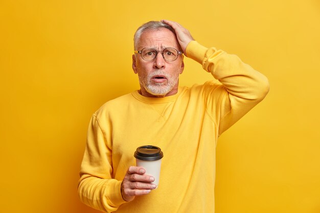 Anciano barbudo avergonzado preocupado mantiene la mano en la cabeza y mira estupefacto el café para llevar de las bebidas delanteras vestido con un jersey casual aislado sobre la pared amarilla no puede creer en el fracaso