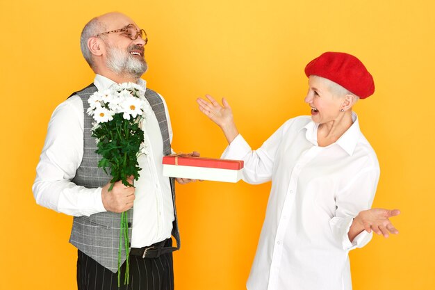 Anciano barbudo con cabeza calva sosteniendo flores de campo y caja de regalo de chocolate para su novia elegante de mediana edad en el día de San Valentín
