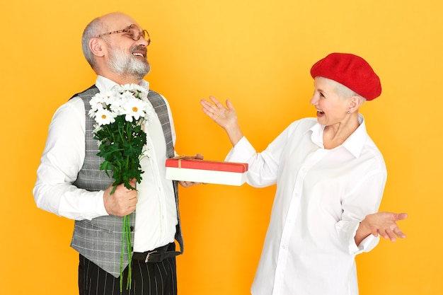 Anciano barbudo con cabeza calva sosteniendo flores de campo y caja de regalo de chocolate para su novia elegante de mediana edad en el día de San Valentín