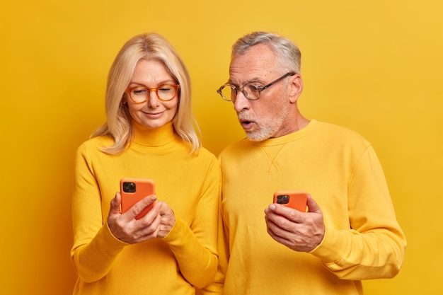 El anciano barbudo conmocionado mira fijamente el dispositivo de teléfono inteligente moderno que muestra que la esposa usa aparatos modernos y lee noticias impactantes en Internet aisladas sobre una pared amarilla