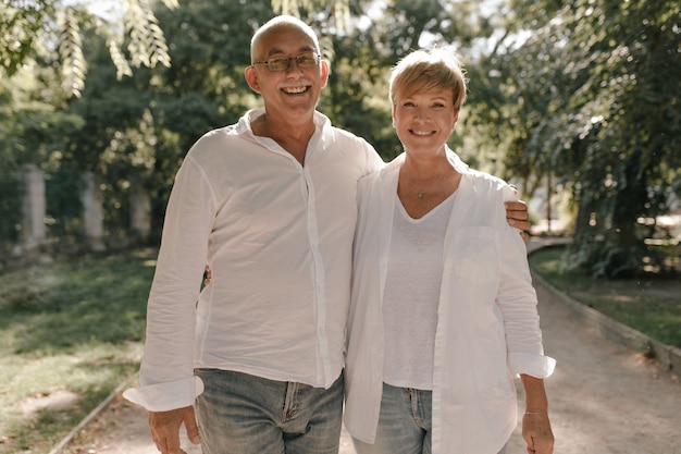 Anciano con bigote gris y anteojos en camisa blanca elegante y jeans abrazando a su esposa sonriente con cabello rubio en blusa ligera en el parque.