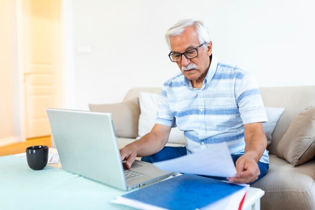 Un anciano de cabello gris enfocado se sienta en el sofá leyendo notificaciones bancarias calculando los gastos domésticos concentrados hombres maduros modernos consideran el papeleo financiero pagan facturas en una computadora portátil en línea