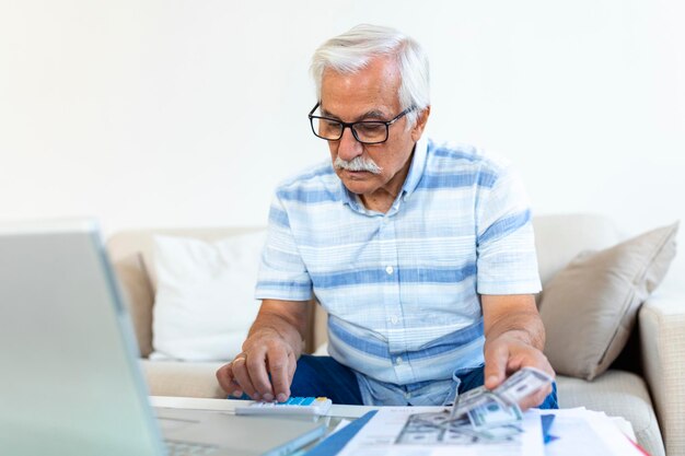 Un anciano de cabello gris enfocado se sienta en el sofá leyendo notificaciones bancarias calculando los gastos domésticos concentrados hombres maduros modernos consideran el papeleo financiero pagan facturas en una computadora portátil en línea