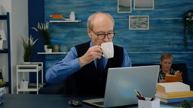 Anciano cansado sentado en el escritorio y tomando un sorbo de café, mirando la computadora portátil trabajando desde el espacio de trabajo en casa mientras su esposa está leyendo un libro en segundo plano. Viejo empresario centrado comprobando gráficos