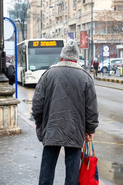 Foto gratuita un anciano con una chaqueta y un sombrero de lana en una parada, calle al fondo, tiempo nublado en bucarest, rumania