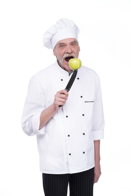 Anciano, chef profesional en uniforme blanco sosteniendo un cuchillo de metal con manzana verde mientras permanece en la pared blanca