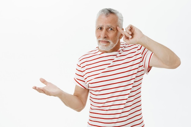 Anciano cosas adolescentes perdieron la cabeza locos Confundido y disgustado anciano barbudo en camiseta a rayas rodando el dedo en la sien por decepción levantando las palmas sin idea reaccionar a la acción tonta