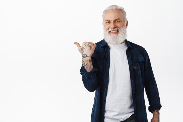 Anciano elegante, chico hipster senior con barba y tatuajes apuntando con el dedo hacia la izquierda, mostrando la promoción a un lado, demostrando publicidad en el espacio de la copia en blanco, pared del estudio