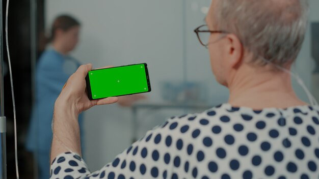 Anciano enfermo que sostiene el dispositivo de la pantalla verde en la instalación en la sala del hospital para el pa ...