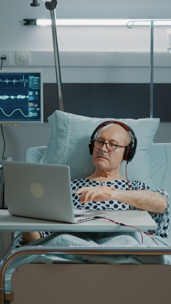 Anciano enfermo sentado en la sala del hospital con ordenador portátil y auriculares escuchando música. Paciente con enfermedad que se relaja mientras recibe tratamiento médico con una bolsa de goteo intravenosa y un tubo de oxígeno nasal