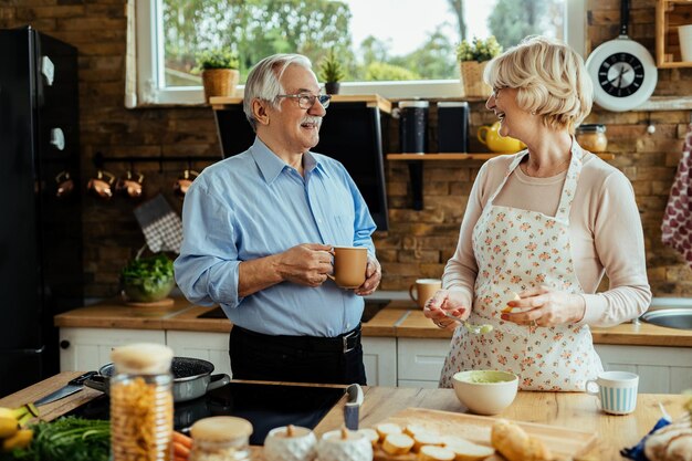 Un anciano feliz bebiendo café y comunicándose con su esposa que está cocinando en la cocina