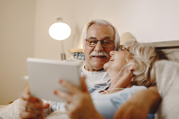 Un anciano feliz y su esposa se divierten mientras usan una tableta digital y se relajan en la cama