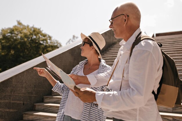 Anciano con gafas y ropa ligera con cámara y mochila sosteniendo un mapa y mirando hacia otro lado con una mujer con sombrero, blusa azul y camiseta blanca al aire libre
