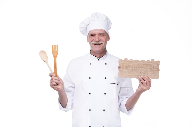 Anciano jefe en uniforme de cocinero sonriendo mientras sostiene el plato de bienvenida, la cuchara y el tenedor aislado sobre la pared blanca
