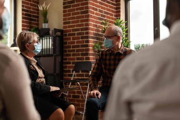 Anciano con mascarilla hablando con pacientes en círculo en una reunión, para curar la adicción al alcohol en la terapia de grupo de apoyo. Hombre discutiendo con psicólogos y personas sobre problemas.