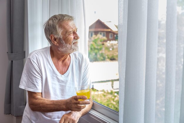 Anciano mayor de pie y mirando por la ventana en el dormitorio después de despertarse por la mañana con jugo