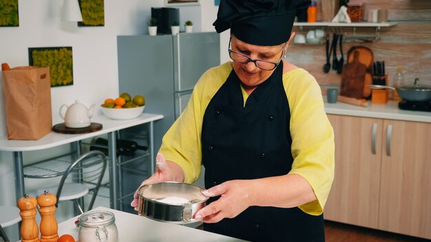 Anciano panadero añadiendo harina en un colador metálico y espolvoreando la masa. Feliz chef anciano con uniforme de cocina mezclando, rociando, agregando, tamizando extendiendo los ingredientes crudos para hornear pan tradicional