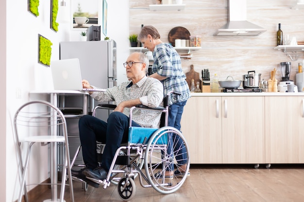 Anciano paralítico en silla de ruedas escribiendo en la computadora portátil trabajando desde casa en la computadora en la cocina mientras la esposa está cocinando el desayuno. Hombre de negocios discapacitado, parálisis de empresario para anciano jubilado