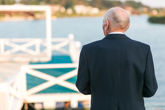 Un anciano de pie a la cámara cerca del muelle en el lago. Vistiendo traje de negocios.