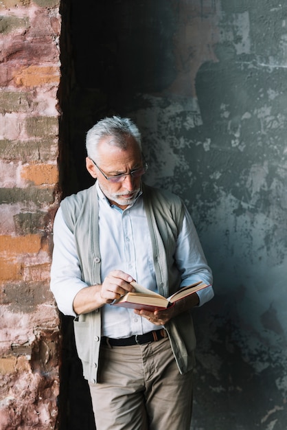 Un anciano de pie frente al muro de hormigón leyendo un libro
