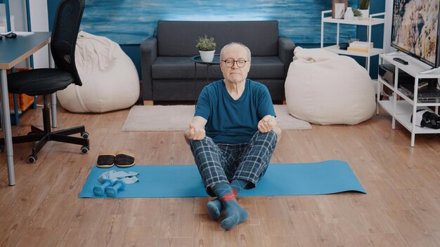 Anciano sentado en posición de loto con los ojos cerrados en la alfombra para practicar la meditación tranquila. Hombre mayor que usa pose de yoga para meditar y encontrar un equilibrio relajante. Pensionista haciendo actividad de bienestar