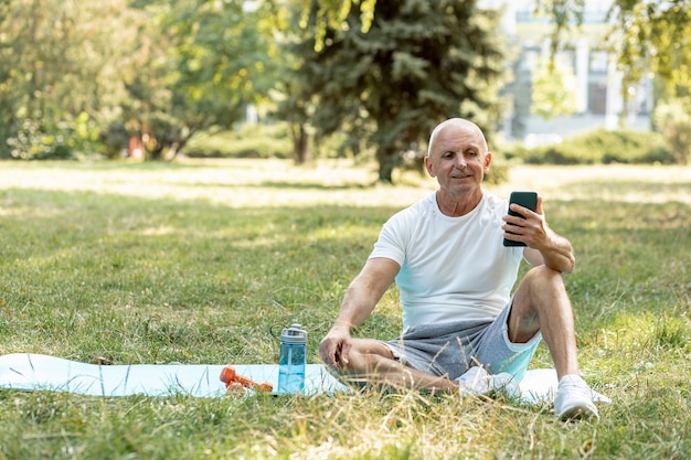 Anciano sonriente checando su teléfono con colchoneta de yoga