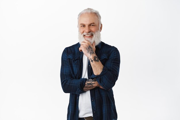 Un anciano sonriente tocándose la barba y mirando complacido a la cámara pensando en escuchar una propuesta interesante asintiendo con aprobación de pie contra el fondo blanco