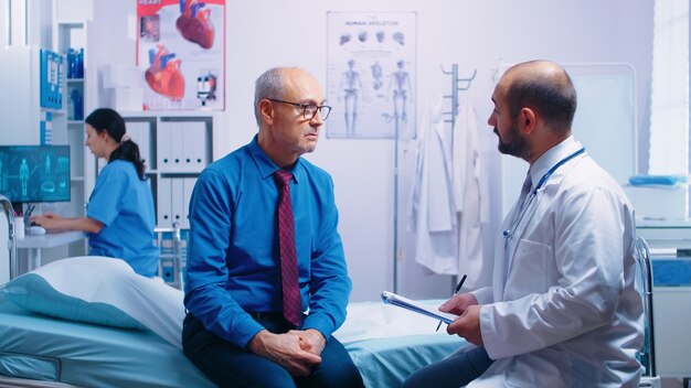 Ancianos jubilados en clínica privada moderna respondiendo al cuestionario médico sentado en la cama de un hospital. Paciente enfermo que busca asesoramiento médico para la prevención de enfermedades del médico generalista en pri moderno