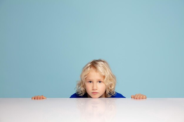 Foto gratuita Ángel. muchacho rizado feliz aislado en el fondo azul del estudio. se ve feliz, alegre. copyspace para el anuncio. infancia, educación, emociones, concepto de expresión facial. asomándose desde detrás de la mesa