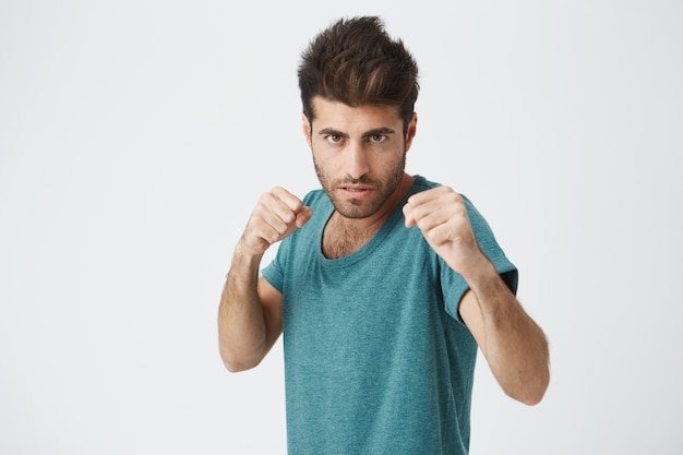 Angry apuesto joven español sin afeitar en camiseta azul de moda, corte de pelo de moda, expresión concentrada, entrenamiento con saco de boxeo para competiciones de boxeo.