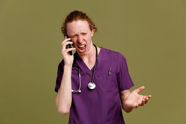 Angry habla por teléfono joven médico masculino vistiendo uniforme con estetoscopio aislado sobre fondo verde