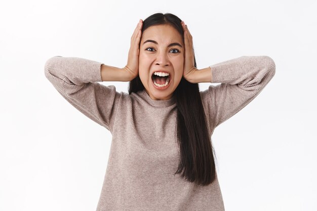 Angustiada y harta, deprimida, alarmada, mujer furiosa, perdiendo el control, sufriendo un colapso mental, gritando agarrar la cabeza y mirar a la cámara con dolor e ira, gritar furiosa, pararse en la pared blanca