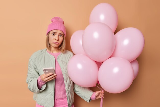 Angustiada joven infeliz vestida con un sombrero rosa chaqueta usa el teléfono móvil para enviar mensajes de texto sostiene un montón de globos inflados celebra cumpleaños aislado sobre fondo de estudio beige