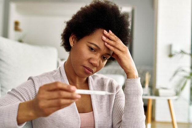 Angustiada mujer afroamericana usando termómetro y controlando su temperatura en casa