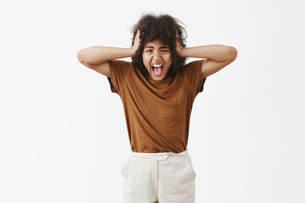 Angustiado cabreado harto molesto adolescente con corte de pelo afro gritando tomados de la mano en la cabeza siendo infeliz con los padres discutiendo cada vez gritando de sentimientos negativos sobre la pared gris, perdiendo la cabeza