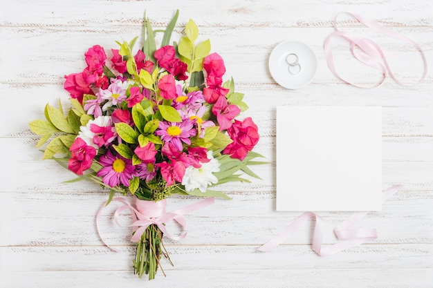 Anillos de boda; Cinta y ramo de flores junto a la tarjeta blanca en el escritorio de madera