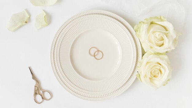 Anillos de boda en placa de cerámica con rosas y tijera sobre fondo blanco