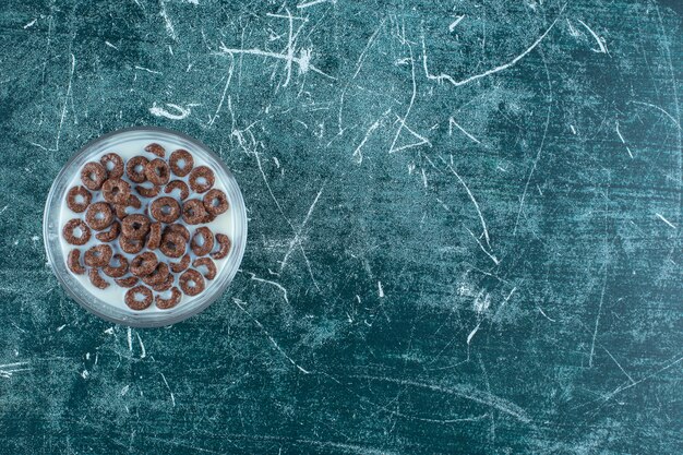 Anillos de copos de cereal de chocolate con leche en un recipiente de vidrio sobre la superficie azul