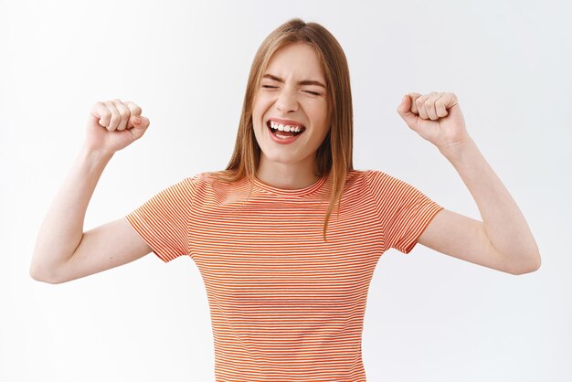 Animada, alegre y guapa mujer caucásica con camiseta a rayas enraizando y gritando de asombro y alegría sintiendo libertad y felicidad puñetazos con una sonrisa y los ojos cerrados triunfando