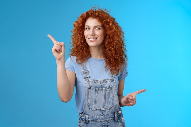 Animado, alegre, guapo, pelirroja de 25 años, pelirroja, de pelo rizado, descarado, verano, estado de ánimo positivo, mono de baile, apuntando hacia los lados con los dedos índices derechos que dirigen los enlaces de las tiendas de promociones, sonriendo encantado.