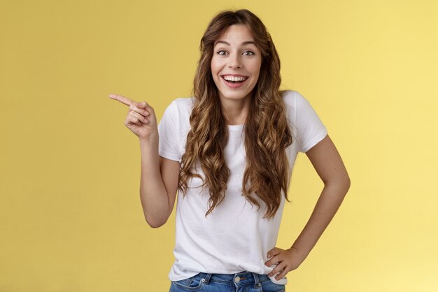 Animado sorprendido feliz entusiasta buena mujer peinado largo y rizado camiseta blanca riendo impresionado asombrado apuntando con el dedo índice izquierdo discutir interesante exposición fondo amarillo