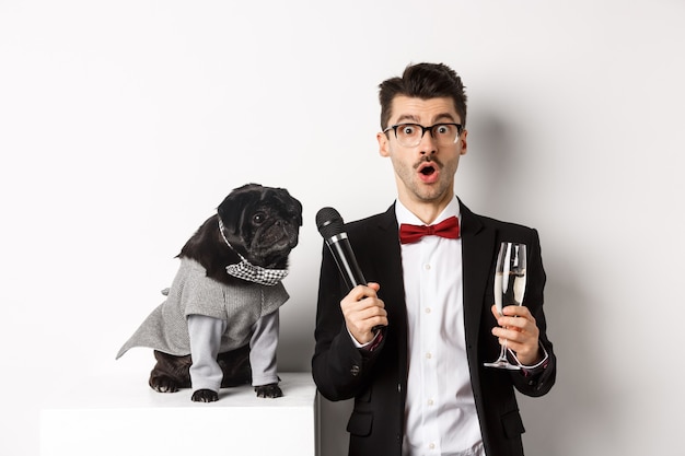 Animador masculino levantando una copa de champán, dando el micrófono al lindo perro negro, de pie sobre fondo blanco.